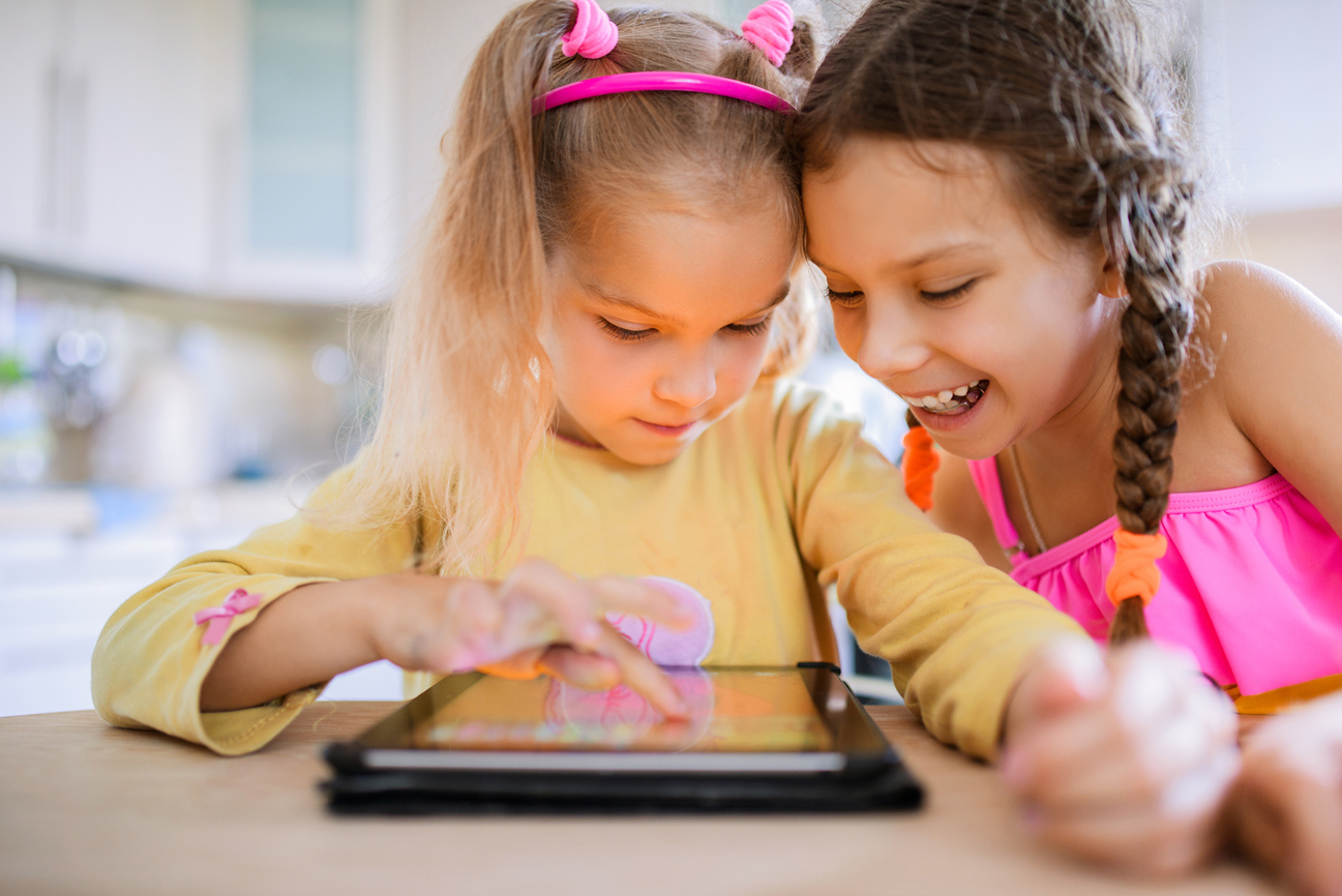 Two beautiful little sisters sit at a table and play on a Tablet PC.